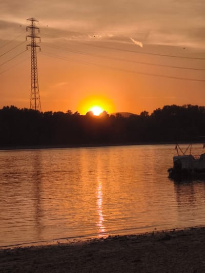 This media file depicts a serene sunset over a body of water, likely a river or lake, in Dunakeszi, Hungary. The sun is partially obscured by trees and hills on the horizon, casting a warm, orange glow across the sky and water. The sky is a soft orange, with wispy clouds and a contrail from an airplane visible in the upper portion. A tall, lattice-style electrical transmission tower stands prominently on the left side of the frame, silhouetted against the bright sky. Power lines extend horizontally from the tower across the image.
The water's surface is calm, with gentle ripples reflecting the vibrant colors of the sunset. A small stretch of sandy shore with scattered pebbles is visible in the foreground. In the distance on the right side of the water, a portion of a boat, possibly a pontoon boat, is docked.
The overall mood is peaceful and tranquil, characteristic of a sunset. The warm, inviting colors suggest a pleasant end to the day. The presence of the power tower indicates a human-influenced landscape, contrasting with the natural beauty of the sunset. No text is visible in the image. The time of day is clearly sunset. The weather appears clear apart from the clouds.
