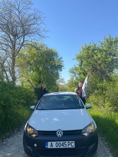 The image shows two people in a white Volkswagen car with the license plate "A 0045 PC" in Rish, Bulgaria. The car is parked on a dirt road surrounded by green trees and grass. The sky is blue. One person is leaning out of the driver's side window and the other out of the passenger side, both with their arms raised.