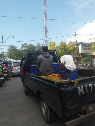 The image shows a black Mitsubishi pickup truck stopped on a street, likely in traffic. Two men are sitting in the bed of the truck, on top of blue and yellow plastic crates. The man on the left is wearing a gray hooded jacket, while the man on the right is wearing a white shirt. A communications tower is visible in the background. Other vehicles, including a silver minivan and motorcycles, are present on the street. A sign for "Restoran CIPANAS" is visible on the right side of the frame. The location is not Khe Luang, Laos.