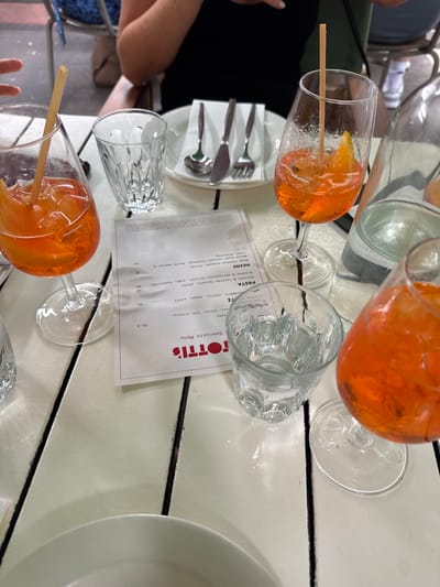 A restaurant table setting includes multiple glasses, some filled with orange cocktails and adorned with straws. A menu for "Totti's Specials Menu" is visible, listing items such as "Clamari," "Spinach & ricotta ravioli," "Brisket & mortadella ravioli," "Wood roasted snapper Fillet," and "Wood roasted Flathead," along with prices. Silverware is neatly arranged on a plate. The table surface is white, with dark dividing lines.