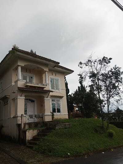 The media file displays a two-story house with a cream-colored facade and a brown tiled roof, situated on a gentle slope next to a paved road. The house features a prominent upper balcony with white columns and railings, and several white-framed windows on both floors. Below the upper balcony is a covered entrance on the ground floor with a white door, flanked by more windows. An outdoor light fixture is visible on the left side of the house's ground floor.
To the right of the house, a grassy mound slopes upwards, dotted with scattered vegetation. Concrete steps lead up this mound towards the house's main entrance, with a simple metal railing at the bottom. Beyond the house and the grassy area, several trees are visible, including one with bare branches and another with dense foliage, possibly coniferous trees. The sky is uniformly overcast, indicating a cloudy day with muted lighting. There are no people or discernible activities taking place in the image. No visible text is present. The scene depicts a quiet residential setting in Amaldiha, India.