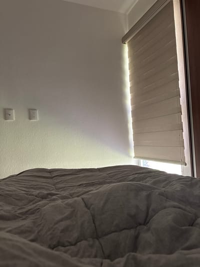 The image displays an interior room, located in Atizapán de Zaragoza, Mexico. The lower half of the frame is occupied by a rumpled, light grey quilted comforter, consistent with being on a bed. In the mid-ground to the left, a plain, light-colored, textured wall features two white, recessed electrical outlets and a smaller, rectangular white wall plate to their left, indicative of a light switch. To the right, a window is partially covered by a beige zebra blind, which is lowered. A prominent vertical shaft of bright natural sunlight penetrates the room through the gap between the blind and the right wall/window frame, casting a strong horizontal band of light across the lower portion of the left wall. A dark brown vertical element, appearing to be a door frame or furniture edge, is visible on the far right side of the frame. No individuals are present in the scene.