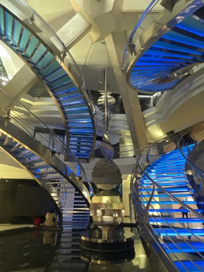 The image shows an interior view of a spiraling staircase in a building, likely a public space, in Les Escaldes, Andorra. The staircase features metal railings with vertical blue light strips. The steps have a glass or translucent material. The architecture of the building is modern with rounded white support structures. There are reflections visible on the polished surfaces of the stairs. A person is on one of the lower levels of the staircase, dressed in a white shirt and a red sweater.