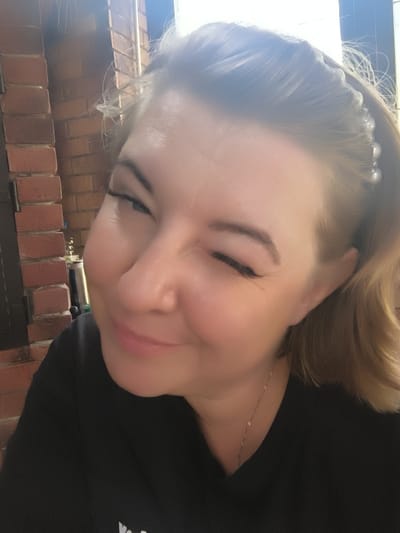 A close-up, slightly overexposed selfie shows a fair-skinned woman with blonde hair styled with a pearl headband. She is winking, with one eye closed and the other looking towards the camera. Her makeup includes winged eyeliner. She is wearing a black t-shirt and a thin gold necklace. The background features a red brick wall and a window with bright light filtering through. The woman is positioned in what appears to be an indoor setting, possibly a home, in Востец Снт, Russia. The lighting suggests it is daytime.