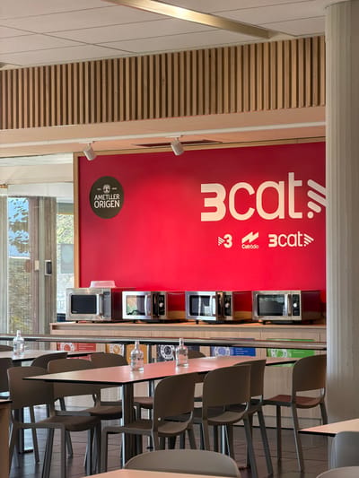The image captures an interior scene, likely a communal eating area or break room, within the Corporació Catalana de Mitjans Audiovisuals (CCMA) building in Sant Joan Despí, Spain. The prominent feature is a vibrant red wall adorned with the "3CAT" logo in large white lettering, accompanied by smaller logos for "3" and "Catràdio." Below the wall, a row of several silver microwave ovens is displayed on a counter.
To the left of the red wall, a circular emblem with "AMETLLER ORIGEN" and "EST. 1830" is visible. The environment includes tables and chairs, suggesting a functional space for dining or relaxation. In the background, tall, vertical wooden slats form a decorative element on the upper wall. The lighting appears to be artificial, with spotlights visible on the ceiling. The scene is devoid of people, with only tables, chairs, appliances, and wall graphics.