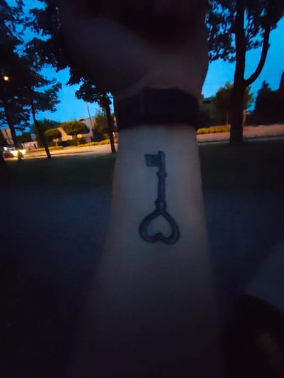 The image shows a close-up of a tattoo of a key on a person's forearm. The key has a heart-shaped bow and an ornate shaft. The person is wearing a dark wristband above the tattoo. The background is blurred and appears to be an outdoor setting at dusk or night, with trees, streetlights, and buildings visible. The location is identified as Stryków, Poland.