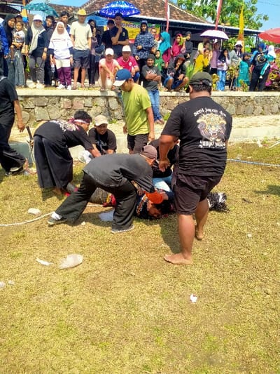 The image depicts an outdoor event in Celep, Indonesia, under bright, direct sunlight.
In the foreground, on a patchy grassy area with visible dry earth and scattered small pieces of white debris, a man is lying prone, face down. He is dressed in dark clothing, with orange and red fabric visible around his head and neck, possibly part of an elaborate costume. Three other men are actively interacting with the prone individual. One man, wearing black pants, a black shirt, and a red headband, is bent over him from the left. Another man, also in black pants and a black shirt featuring a white design and the text "Seni Jaran Dwi Setyo Mudo" on the back, is bent over the prone man from the center. To the right, a third man, barefoot, wearing black shorts and a black t-shirt with the same "Seni Jaran Dwi Setyo Mudo" text and a tiger/demon head graphic, stands looking down, his hands placed on the back of the second bent-over man. A fifth man, wearing a lime green t-shirt and light-colored shorts, stands slightly behind and to the left of the main group, observing. A sixth man, wearing a dark t-shirt and a light-colored cap, is seated on the ground behind the central action, watching. A white rope lies on the ground to the far left.
In the midground, a crowd of approximately 20-30 spectators, comprising adults and children in various casual attire, observes the scene from behind a low stone or concrete wall. Several spectators are holding assorted umbrellas (blue, green, pink, white) for shade. In the background, signs with Indonesian text, including "WARUNG PECEL BE..." and a banner with red and white stripes, are visible. Buildings with tiled roofs and some green foliage are also present, beneath a clear blue sky.