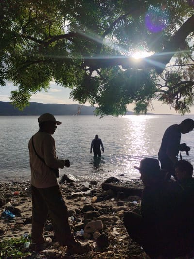 The image shows a coastal scene in Lamreh, Indonesia. In the foreground, a man wearing a cap and casual clothes stands on a rocky beach littered with trash. He is silhouetted against the bright sunlight reflecting off the water. To his right, a person is waist-deep in the water. Further to the right and slightly behind, a man with a camera appears to be photographing. Close to the camera's point of view, two other figures are sitting on the rocky ground, partially obscured by shadows. The background features a bay with hills visible across the water and sunlight is shining through the leaves of a large tree in the upper portion of the frame.