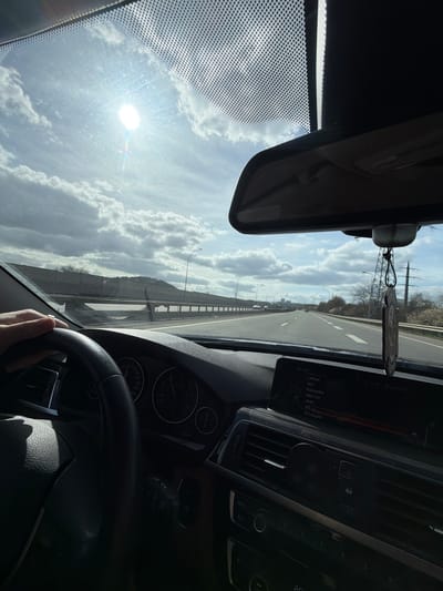 The image, taken from the perspective of the driver inside a car, shows a view of the A13 highway in Dudelange, Luxembourg. The driver's hand is visible on the steering wheel. The dashboard and radio with the station LATINA displayed are visible. The sun glares through the windshield. The highway stretches into the distance, lined with streetlights and a bridge. Above, a cloudy sky fills the frame. A rearview mirror and a hanging object are visible in the upper part of the image.