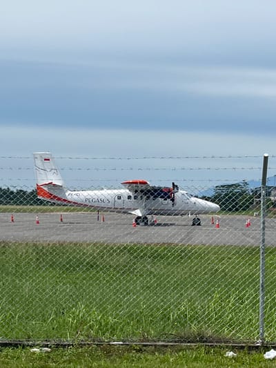 A twin-engine propeller aircraft with the registration PK-ICI and the word "PEGASUS" written on its fuselage is parked on an asphalt tarmac, as seen through a chain-link fence. The aircraft is white with orange accents on its tail and wings. The tail fin displays the Indonesian flag. Several orange traffic cones are positioned around the aircraft. The scene is set against a backdrop of green grass, trees, and a cloudy sky. This image was taken in Peurupok, Indonesia.
