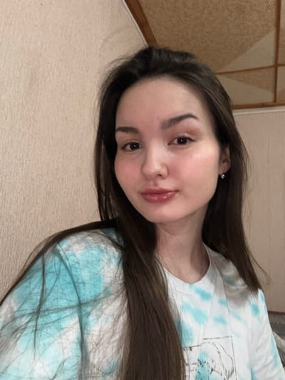 The image depicts a young adult female in an indoor setting located in the city of Bayronovka, Russia. The female, with long, dark brown hair and light complexion, is captured in a front-facing portrait. She wears a white t-shirt featuring a light blue tie-dye pattern and a partial dark graphic design on the lower right. Her right ear displays a silver-colored stud earring with a clear stone and a smaller stud in the cartilage. Her lips are full and glossy, with a slight smile. The background consists of a textured, light beige wall on the left and a wooden ceiling beam or trim element visible along the top edge, leading to a lighter, possibly paneled ceiling section on the upper right. Illumination appears to be from a diffuse, warm indoor light source.