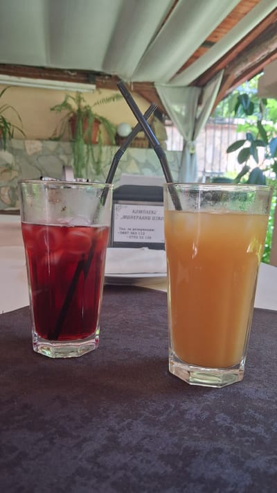Two glasses of beverages sit on a dark textured surface. The glass on the left contains a deep red liquid with ice and a black straw. The glass on the right contains an orange-colored liquid with ice and a black straw. Both straws are bent and crossed. In the background, a sign with Bulgarian text is visible, which reads "КОМПЛЕКС," "МИНЕРАЛНИ ИЗВОРИ," and phone numbers. The setting appears to be an outdoor covered area, possibly a restaurant or cafe, with draped white fabric overhead and some greenery and stone walls visible in the background. The overall ambiance suggests a relaxed, outdoor dining experience. This location is identified as being in the city of Marchevo, Bulgaria.
