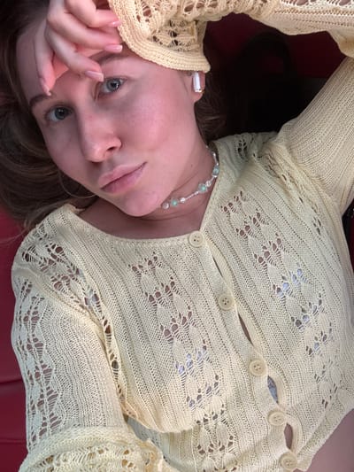 A woman, seen from a high angle, lies on a red surface, possibly a car seat. She wears a yellow, knitted, button-up cardigan with an intricate open-knit pattern. A delicate pearl and green bead necklace adorns her neck. Her right hand is raised to her forehead. She also wears white wireless earbuds. The lighting is warm, and the image appears to be a selfie. No specific location in Fuyang, China, is identifiable.