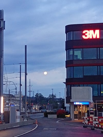 The image depicts a view of the 3M building in Langenthal, Switzerland, at dusk. The sky is a deep blue, with a full moon visible in the upper left quadrant. The building, a modern multi-story structure with a curved facade, is illuminated by red neon "3M" lettering on its upper section. The lower part of the building features large, reflective windows. In the foreground, a paved road curves around a concrete barrier, with a single orange traffic cone on the left side. Construction barriers with red and white stripes are visible to the right, indicating ongoing work. In the middle ground, there are railway tracks and overhead power lines, suggesting the proximity of a train station or transportation hub. A small, illuminated structure, possibly an entrance or information kiosk, is situated at the base of the 3M building, and a dark-colored car is parked in the background to the left of this structure. Lush red foliage is planted in front of this entrance. The overall scene is one of an urban environment at twilight, with a blend of corporate architecture and infrastructure.