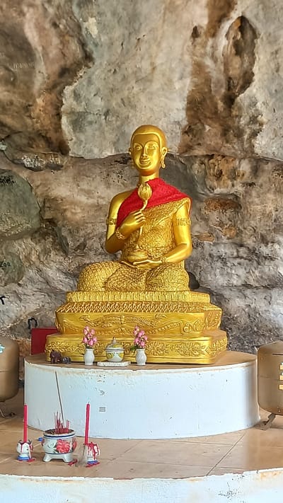 A gold Buddha statue sits on a platform in a cave in Thap Put, Thailand. The statue is adorned with a red sash and holds a flower. Offerings of flowers, incense, and candles are arranged on the platform in front of the statue. The cave walls are rough and textured.