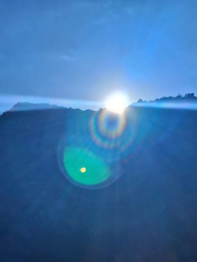 The image captures a dramatic sunset over a mountainous landscape, likely near Vayk, Armenia. The sun, a brilliant orb of white light, is setting behind a silhouetted mountain range, its rays creating a strong lens flare that dominates the center of the frame. The flare is characterized by concentric circles of light in various colors, including yellow, green, blue, and pink, emanating from the sun. The sky above is a deep, clear blue, with hints of wispy clouds. The foreground is a dark, shadowy mountain slope, largely obscured by the low light. The overall impression is one of a powerful natural spectacle, with the lens flare adding an almost ethereal quality to the scene.