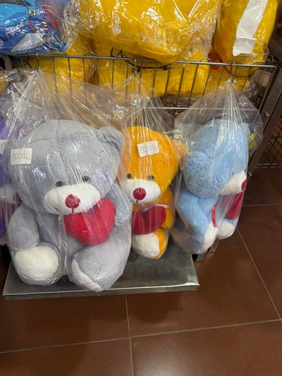 Three teddy bears, each approximately 12-15 inches tall, are displayed on a metal shelf. The teddy bear on the left is gray, the middle one is orange, and the one on the right is blue. All three bears are holding a red heart. Each bear is individually wrapped in clear plastic, and each has a white price tag with "800 TL" written on it. The bears are positioned in front of a rack of yellow plush items, possibly pillows or blankets, also wrapped in plastic. The background consists of a wire rack and the floor is tiled in a reddish-brown color. The image appears to be taken inside a store. Acıpayam is a town and district in the Denizli Province of Turkey.