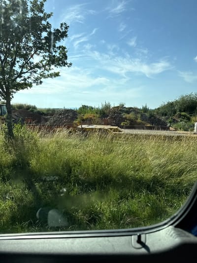 The image shows a view from inside a vehicle, looking out at a grassy field and a construction site. The sky is a light blue with wispy white clouds, suggesting a clear day. The time of day appears to be afternoon, as indicated by the angle of the shadows.
In the foreground, to the left, a portion of a green tree with dark leaves is visible, its branches reaching towards the sky. The main subject of the image is a landscape that appears to be undergoing development. A grassy embankment slopes upwards, with a light-colored railing or fence running along its top. Below the railing, there's a darker area with exposed earth and some construction materials. A yellow piece of construction equipment, possibly a loader or excavator, is partially visible on the site.
The right side of the image shows the interior of the vehicle, specifically the side of the window frame, which is dark gray and reflective. The reflection in the glass shows more of the green grass and sky. The overall scene suggests a rural or semi-rural setting in Oberwetz, Germany, possibly a construction project near a natural landscape. No people are clearly visible in the image. There is no discernible text in the image.