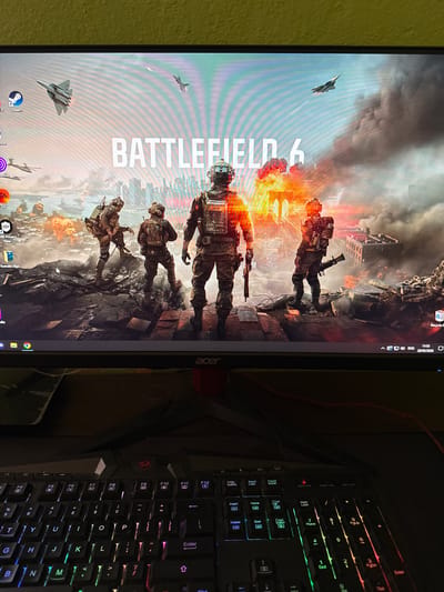 The image depicts a computer screen showing a Battlefield 6 game wallpaper. The wallpaper features soldiers in a war-torn environment with fighter jets flying overhead. Below the screen are visible icons for Steam, a browser, and the Electronic Arts application. To the right of the screen is a black RGB gaming keyboard. The wall to the left of the screen is painted in a muted yellow or light green color. The time and date, "11:53 29/05/2026", are visible in the top right corner of the screen. The location in the city of Bratya Daskalovi, Bulgaria cannot be determined from the image.