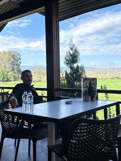 An adult male is seated at an outdoor table on a patio, which overlooks a rural landscape in Ula, Turkey. The man, estimated to be in his 40s or 50s with a short beard, wears a dark long-sleeved shirt and is positioned on the left side of the frame, looking towards the camera with a subtle expression. He holds a lit cigarette in his right hand. On the dark-surfaced table in front of him are two transparent plastic bottles of Damla brand water, a silver-colored round ashtray containing ash and a cigarette stub, and a menu stand displaying text in Turkish ("RAMAZAN'A ÖZEL") alongside food images. Adjacent to the menu are two stainless steel shakers and a dark bottle. The seating consists of dark, open-weave plastic chairs. The patio structure includes dark brown pillars and a slatted ceiling. Beyond a dark railing, the environment consists of expansive green fields, scattered trees (some deciduous, some coniferous), and distant mountains under a blue sky with scattered white clouds. A prominent conifer tree stands in the immediate outdoor view.