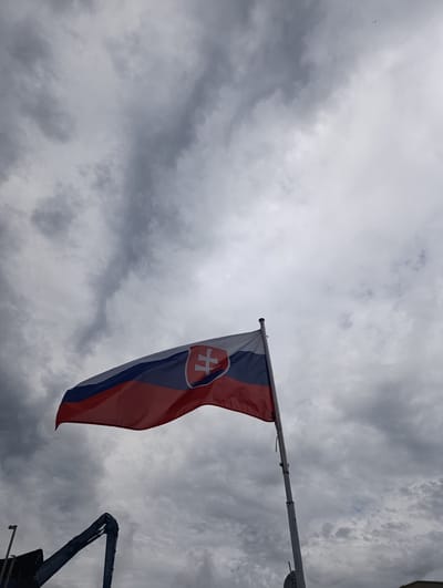 The image depicts the flag of Slovakia, identified by its tricolor design of white, blue, and red horizontal stripes, with a coat of arms placed on the white stripe. The flag is affixed to a pole and is captured mid-flutter against a backdrop of a heavily overcast sky filled with dark, textured clouds. In the lower left portion of the frame, a dark blue mechanical arm, possibly part of a construction vehicle or equipment, is visible, bearing the text "Elixir Zorka." The presence of the "Elixir Zorka" logo suggests the location is likely within or near an industrial area or facility operated by this company, which is known to have operations in Šabac, Serbia. The scene is devoid of any visible people or human interaction.