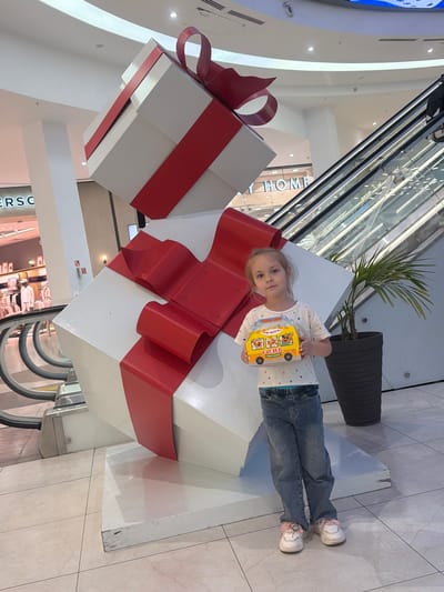 A young girl stands next to a large decorative stack of white gift boxes wrapped with wide red ribbons and bows, seemingly within a shopping mall. The girl is holding a yellow toy bus. She is wearing blue jeans, a white shirt with small hearts, and pink and white sneakers. The mall interior features an escalator behind the girl and the gift box display. A plant in a grey pot sits nearby. The tiled floor suggests a public space within the mall. The location is Novoaleksandrovka, Russia.