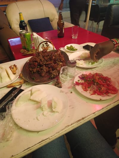 The image captures a casual dining scene, likely in a restaurant or bar, characterized by a vibrant mix of food and beverages, suggesting an evening or nighttime gathering.

In the foreground, a distressed white wooden table is visible, with a person's legs in blue jeans positioned underneath. On this table, there is a white plate holding several irregularly shaped, chunky pieces of white food, strongly resembling ugali, a stiff porridge common in East Africa. To the left of this plate, a knife with a dark handle rests on the table, next to a crumpled clear plastic bag or wrap. Further to the right on the white table, another white plate contains a colorful salad composed of diced red tomatoes and thinly sliced red onions, possibly mixed with some green herbs or chili, known as kachumbari.

Dominating the mid-ground is a bright red, glossy table laden with various items. At its center, a large, round metal platter is piled high with numerous pieces of dark, cooked meat, consistent with grilled or fried goat meat (nyama choma), a popular East African dish. Around this platter, several other objects are arranged. On the left side of the red table, a green soda can with an orange label is visible, along with a green bottle with a yellow cap and a blue label (possibly a beer like Tusker). A pile of white folded napkins rests in a holder decorated with a circular design featuring an animal, possibly an antelope or deer.

On the right side of the red table, a brown beer bottle and a clear empty glass are placed. Closer to the viewer, next to the meat platter, are a salt shaker and another shaker (possibly pepper), alongside a small clear container that appears to hold salt. A small white ceramic decorative item also sits nearby. A white plate holds a small portion of green chili peppers, and another white plate contains a few smaller pieces of ugali. A dark-skinned hand, adorned with a wristband featuring the colors of the Kenyan flag (black, red, green, and white stripes), is reaching towards this plate, seemingly picking up a piece of food. A smartphone is also partially visible on the table.

In the background, partial views of furniture include a beige armchair with a blue cushion on the left. A blurry figure of a person, possibly a patron or staff member, can be seen in the distant background. The overall lighting is dim and artificial, indicating it is not daytime. The presence of specific dishes like nyama choma and ugali, along with the Kenyan flag wristband, strongly suggests an East African, likely Kenyan, setting. The atmosphere appears casual and social, centered around enjoying food and drinks.