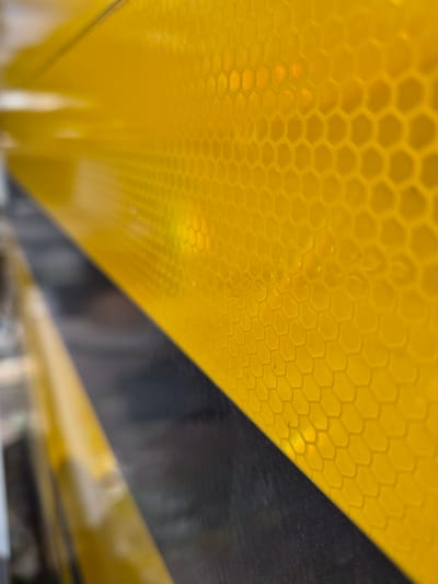 The image is a close-up, macro shot of a yellow, hexagonal, reflective surface, possibly a safety marker or sticker. The surface has a textured, honeycomb pattern. The bottom portion of the image shows a dark, out-of-focus area that appears to be a surface or object below the yellow material, separated by a diagonal line. The lighting suggests the object is outdoors, and there are some reflections or light sources within the hexagonal cells, indicating its reflective property. The image is taken from an angle, giving a sense of depth and texture. The location of Masów, Poland, is not visually represented in this abstract, close-up image.