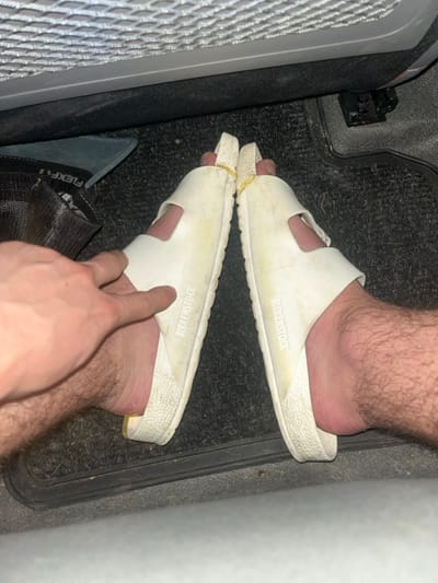 This image depicts a downward view into the interior of a vehicle, located in the city of Azor, Israel. The primary subjects are a person's bare, hairy lower legs and feet, adorned with two white Birkenstock brand EVA sandals. Both sandals display wear, dirt, and discoloration. Yellow rubber bands are present, wrapped around the front strap of each sandal, positioned near the toes. A person's bare, hairy hand, with visible knuckles, is extended into the frame from the left, with the index finger making contact with the inner side of the left sandal. The feet and sandals rest on a dark, textured vehicle floor mat, which shows some visible debris. In the upper portion of the image, a grey mesh fabric, consistent with a vehicle seat-back pocket or storage net, is visible. To the left, a black mesh object, possibly a bag or cap, with partially visible white text "FLEXF", rests on the floor mat. A dark metallic structure, likely a seat rail, is visible on the upper right side of the floor.