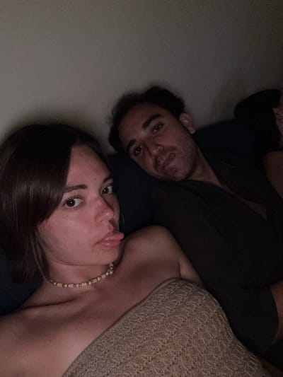 Two individuals, a woman and a man, are captured in a close-up, dimly lit selfie, seemingly taken indoors. The woman, positioned on the left and closer to the camera, has dark brown hair and is looking directly at the lens with her tongue playfully sticking out. She is wearing a strapless top and a beaded necklace. The man, to her right and slightly further back, has dark, curly hair and is looking towards the camera with a relaxed expression. He is wearing a dark shirt. The background is a plain, dark wall. The scene does not provide any specific information about a location in Chevy Chase, United States of America.