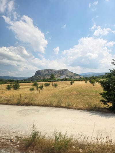 The image depicts a wide, dry, grassy field with scattered bushes and trees under a bright blue sky with scattered white clouds. A flat-topped, rocky hill dominates the background, with a white building visible on its slope. A dirt road or track runs across the foreground. The scene appears to be in a rural area, likely the outskirts of Theopetra, Greece, given the characteristic landscape of the region.