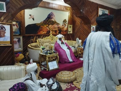 The image shows a room with wooden walls and a large, ornate couch. A man wearing a pink robe and white turban sits on the couch, holding a microphone. A man wearing a white robe and a blue and white turban stands in the foreground, facing the couch. A seated man wearing a white robe and turban sits next to the couch on the floor. A small table with a fan sits in the foreground. There are also two framed portraits on the walls. The scene is in a room located in Kaltungo, Nigeria at the coordinates latitude: 9.816, longitude: 11.315.