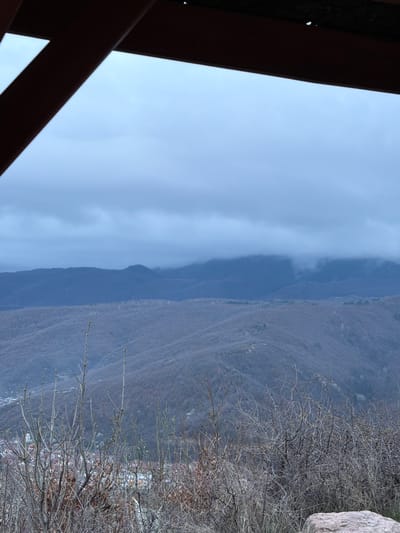 The image shows a landscape view from a building or structure, possibly in the Sveti Konstantin area of Bulgaria. The foreground features the angled wooden supports of a building. Beyond that, the landscape consists of a mountain range shrouded in mist or low clouds. The terrain is rugged, with visible bare trees and foliage in the middle ground. A large rock is visible on the right side. The sky is overcast, contributing to the overall somber tone of the scene. No people are visible.