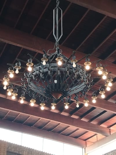 A large, ornate black metal chandelier hangs from a dark brown, paneled ceiling. The chandelier has multiple arms, each supporting a lightbulb. The design features decorative curls and a central, cone-shaped element. A rectangular metal support structure connects the chandelier to the ceiling. Natural light enters from below, illuminating the lower portion of the image.