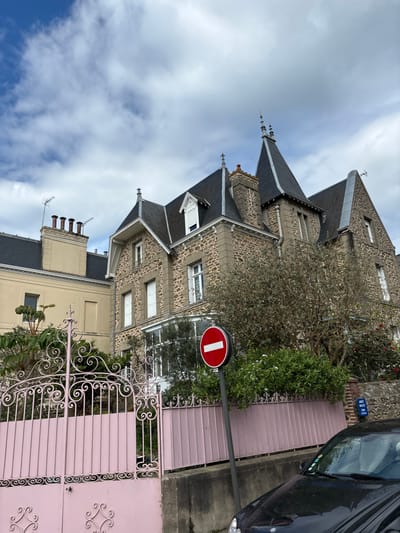 A large, grey, stone, multi-story house with a slate roof, tall turrets, and a decorative iron gate in front. The house is located in a residential neighborhood, with other houses visible in the background. There is a small, green bush to the left of the house. A black car is parked in front of the house on the right side of the image. The sky is blue with white clouds. The street sign says "Rue de la Gare" and a red, white, and blue flag is hanging from a light pole. The stop sign is in the front yard. The scene is likely in a suburban neighborhood of Dinard, France, as the latitude and longitude coordinates indicate a location near the city center.