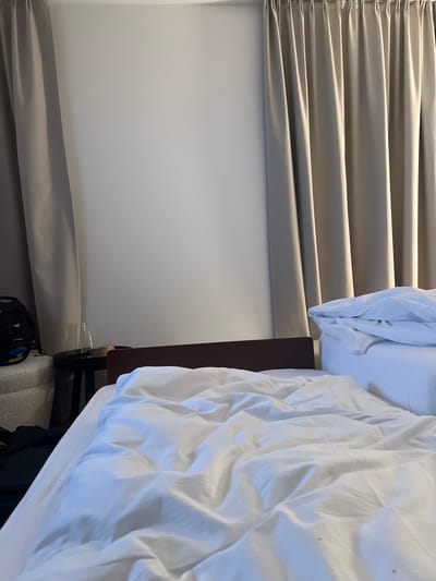 A close-up shot shows part of a hotel room. On the right, a white bed with rumpled bedding spills over the edge. Next to the bed is a dark wood nightstand. To the left, light tan horizontal blinds cover the wall, with the bottom edges forming soft folds. Part of a backpack peeks out from under the bed, adding a touch of personal clutter to the scene.