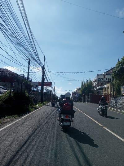 The image captures a sunny daytime street scene in Serpong, Indonesia, as indicated by a prominent "POLISI POLSEK SERPONG" sign on a building to the right. The two-lane asphalt road features a white scooter in the foreground, viewed from behind, carrying a single rider wearing a white helmet and a dark jacket with a red backpack. The scooter's license plate, "5981 FJI", is visible. Ahead in the same lane, a second dark-colored scooter with a rider is present. On the opposite side of the road, another dark scooter with a rider is visible, potentially traveling in the opposite direction. Long shadows from vehicles and roadside objects stretch across the road surface.

To the left, a dense tangle of black overhead utility wires spans across multiple utility poles. Beneath these wires, an overgrown roadside with vegetation and a metal fence borders a building with a reddish-brown tiled roof. Further down, a billboard displaying "MOVILOX" and partially "MECCA" is visible. To the right of the road, a low stone wall with dark, irregular stones supports a two-story building with white walls. This building features a dark gate and pillars, and prominently displays the "POLISI POLSEK SERPONG" sign on its facade. Green foliage and trees are visible around the building. The sky is clear and bright blue with minimal cloud cover.