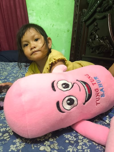 An Asian female child lies on a bed with a blue floral patterned bedsheet. She is wearing a yellow top with ruffled sleeves and a gold chain necklace. To her right, a large pink plush toy with a smiling face and the text "TUNG TUNG SAHUR" embroidered on it is positioned on the bed. The background shows a green wall and a dark wooden carved headboard of a bed. A maroon curtain is visible to the left. The scene appears to be an indoor domestic setting, likely a bedroom. The location is Ujongreuba, Indonesia.
