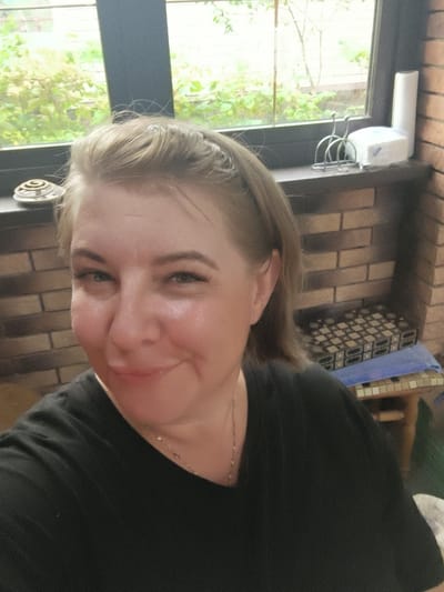 A medium shot photograph, taken from a slightly low angle, features a woman with blonde hair, wearing a black t-shirt, looking directly at the camera with a slight smile. Her face is illuminated, highlighting her features and makeup. She is positioned in the foreground, with a blurred background that suggests an indoor setting with a window. Behind her, a brick wall is visible, and on a ledge near the window, there are several items, including a decorative metal object, a napkin holder, a stack of napkins, and a roll of paper towels. A patterned surface with small squares is also visible in the background, resting on a stool. The overall impression is a casual, personal photograph taken likely in a home or similar private space. The provided location, "Востец Снт, Russia," suggests the setting is a dacha or a cottage in a garden community in Russia.