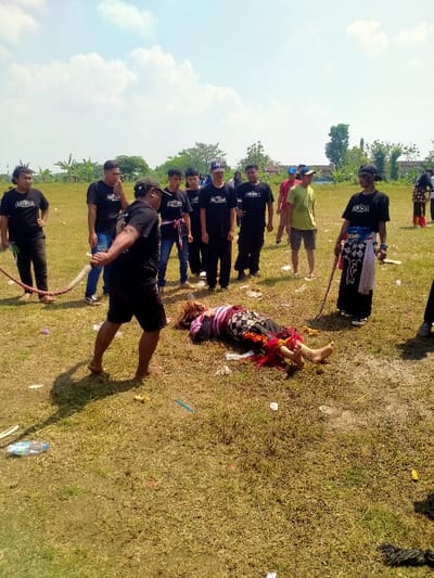 This image depicts an outdoor scene in Celep, Indonesia, on a clear, sunny day in a dry, grassy field.
**Environment:** The setting is an open field with sparse, dry grass and patches of exposed soil. The ground is littered with various discarded items, including plastic bottles, wrappers, and colored debris. In the background, scattered trees, shrubs, and indistinct distant structures are visible under a bright blue sky with sparse white clouds. Strong sunlight creates distinct shadows to the right of the standing figures.
**People and Subjects:** Approximately 12 individuals are present.
* **Central Figure:** A person of indeterminate gender lies prone (face down) on the grass in the center-right foreground. The individual wears a horizontally striped top (dark, pink, white), a dark patterned lower garment (possibly a sarong or skirt), and red fabric with tassels wrapped around the lower legs/ankles. Their feet are bare. The head is turned to the left, and arms appear bent beneath the body.
* **Foreground Left Participant:** An adult male, barefoot and wearing a dark t-shirt and dark shorts, is positioned in the immediate foreground with his back partially to the viewer. He is holding a thick, braided light-colored rope that extends out of frame to the left.
* **Mid-Ground Left Group:** A group of approximately seven adult males stands observing, primarily wearing dark t-shirts (some with logos), some with caps, and some with colorful sashes or belts. One individual in this group also holds the rope.
* **Mid-Ground Right Individuals:** Two adult males are present. One wears a lime green t-shirt and light shorts. The other wears a dark t-shirt, a dark cap, and dark sunglasses, with a checkered black and white sash/skirt over dark pants. This individual holds a thin, dark stick vertically.
* **Background Right Figure:** A partially visible individual in the far right background also appears to be wearing traditional attire, including red fabric, similar to the central prone figure.
**Actions and Interactions:** The central figure is motionless in a prone position on the ground. The surrounding individuals are standing and observing the scene. The rope on the left suggests an ongoing or recently concluded activity, possibly a tug-of-war or ritualistic performance, with multiple participants holding it. No direct physical interaction with the prone individual is evident at the moment captured.