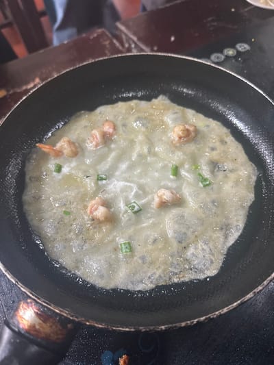 A close-up shot depicts a pan filled with a partially cooked omelet. The omelet is light in color with visible pieces of shrimp and sliced green onions scattered across its surface. The pan itself is black and shows signs of use, with some oil or residue around the edges. The omelet appears to be in the process of cooking, with a slightly wet or glistening appearance.