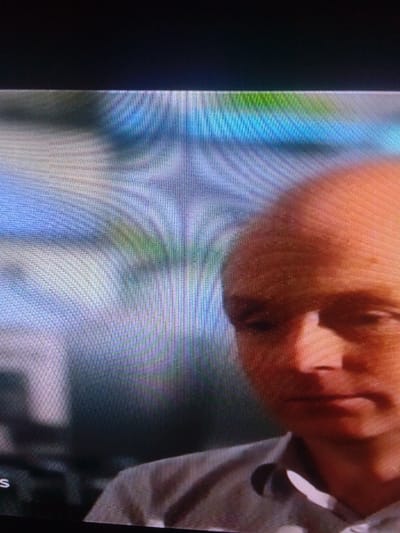 The image presents a photograph of a digital screen, displaying a male individual in a close-up view. The subject, exhibiting a balding or shaven head and light complexion, is visible from the chest up and occupies the right portion of the frame. He is looking downwards and to his left, with a neutral facial expression. The man is dressed in a light-colored collared shirt. The background behind the subject is an out-of-focus, pixelated field of blue, green, and grey colors. A partial white letter 'S' is discernible against a dark background in the lower-left corner of the displayed content. A distinct pixel grid pattern is visible across the entire image, indicating it is a photograph of a screen. The location of this scene is the city of Mista Ali, Nigeria.