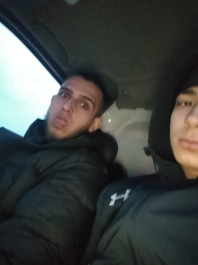 This image captures two males seated within the interior of a vehicle, in what is stated to be Stroevo, Bulgaria. The individual on the left, a male with short dark hair, wears a dark puffer jacket over a dark hooded garment. He displays an exaggerated facial expression with wide eyes and an open mouth, his tongue extended. A small mark is visible above his nose. The second individual, partially visible on the right, is a male wearing a dark hooded sweatshirt featuring a white 'H' logo on the left chest. His face is partially obscured by the hood and the image frame, presenting a neutral expression. The vehicle's interior includes a dark ceiling and a light-colored panel in the background. A window frame in the upper left corner reveals a bright, undifferentiated exterior. The scene is illuminated by ambient light, appearing dim.