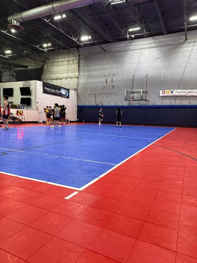 The image shows an indoor volleyball court in Rockville, Maryland. The court has red and blue playing surfaces. There is a group of volleyball players standing on the blue area of the court.  A basketball hoop is visible in the background, along with banners for DMV Volleyball Academy and Vienna Elite Volleyball Club. A scoreboard and other equipment are visible on the side of the court. The ceiling has visible metal supports and lighting.