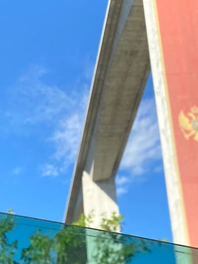 The image shows a view of the Millenium Bridge in Gornje Mrke, Montenegro, under a clear blue sky with sparse white clouds. The bridge's concrete structure dominates the upper portion of the frame, arching diagonally across the sky. To the right of the bridge, a tall, red structure with a golden emblem is visible, appearing to be part of a flag or banner. In the foreground, a blue-tinted reflective surface, likely glass, runs horizontally across the bottom of the image, partially obscuring the view of green foliage beneath it. The scene is captured from a low angle, emphasizing the bridge's scale against the sky.