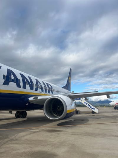 A Ryanair Boeing 737 aircraft is parked on the concrete tarmac of Girona-Costa Brava Airport in Vilobí d'Onyar, Spain, under a partially overcast sky. The left side of the aircraft dominates the foreground, featuring a white fuselage with a prominent blue "RYANAIR" logotype and a yellow stripe running beneath the window line. The aircraft's registration, "EI-DCH," is visible on the rear fuselage. The left engine nacelle is detailed, with its internal fan blades clearly visible. A set of white mobile stairs, also branded "RYANAIR," is positioned at the aircraft's rear passenger door. In the background, another aircraft, identifiable by its red and white livery as a Jet2.com plane, is partially visible. A solitary individual wearing a yellow high-visibility vest stands on the tarmac near the Jet2.com aircraft, with a partial view of a black ground service vehicle behind them. Distant mountains form the horizon line beneath the cloudy sky.