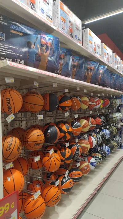 The image shows shelves in a retail store displaying basketballs and basketball-related toys. The shelves are stocked with a variety of basketballs in different colors, sizes, and brands, including Molten and Spalding. Some of the basketballs are orange, while others have black, white, and colored patterns. There are also boxes on the upper shelves containing basketball game sets, such as a "Basketball Double Shot Light-Up" game. The store appears to be a toy or sporting goods store. The scene is brightly lit and organized, suggesting a well-maintained retail environment. The overall impression is that of a commercial setting where sports equipment for children is being sold.