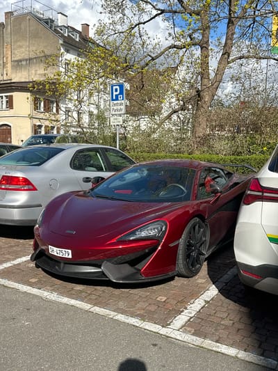 The image shows a red McLaren sports car parked between two other vehicles in a designated parking area. The area is identified by a blue and white parking sign that reads "Zentrale Parkuhr, ZONE 1 max. 2 Std," indicating a central parking area with a maximum two-hour limit. The license plate of the McLaren is "SH-47579." The parking area is paved with dark bricks and delineated by white painted lines. A building with visible architectural details is visible in the background, along with tree branches in front of it. The OCR labels "BACK PACKER" and "201" can be seen on the building. The photo was likely taken in the city of Schaffhausen, Switzerland, given the license plate and sign.