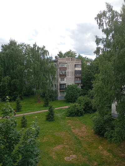 This is an outdoor, eye-level shot of a residential building in Gorki-2, Russia. The building, constructed of concrete panels, features multiple floors with balconies and windows. It is surrounded by lush green trees and grass. A paved walkway runs through the grassy area. A person wearing an orange jacket and dark pants is walking on the path. The sky is overcast with clouds.