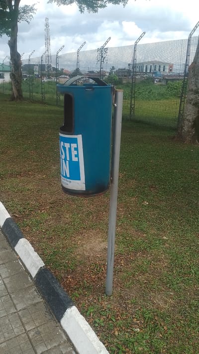 A blue metal waste bin stands attached to a gray metal post on a grassy lawn. The bin has a white rectangular sticker with the words "WASTE BIN" printed on it. The curb of a gray-tiled walkway runs along the edge of the grass. The background features a tall metal fence with what appear to be security spikes. Beyond the fence are trees, buildings, and a communication tower in the distance. The sky is overcast with gray clouds. The location is Asubi, Nigeria.